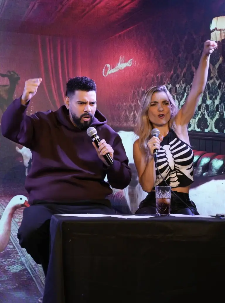 Dos artistas sentados a una mesa en el escenario, uno levantando un brazo