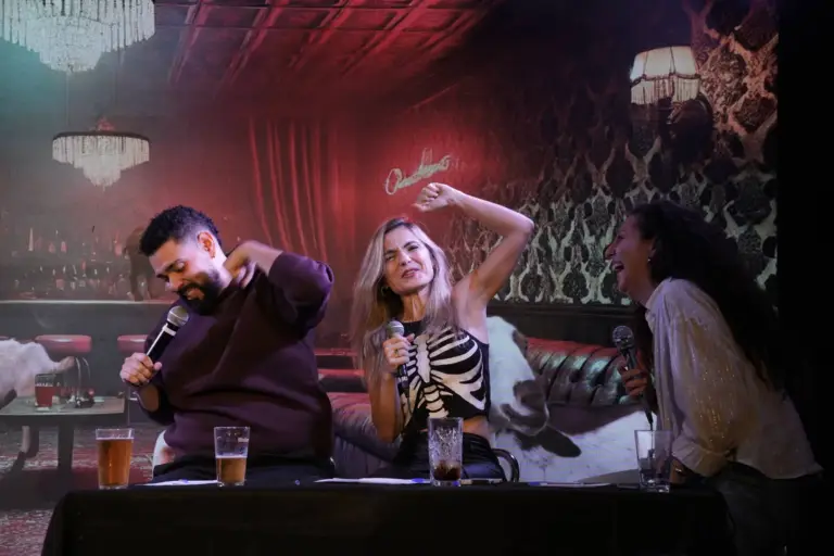 Tres artistas a una mesa en el escenario reaccionando a un chiste