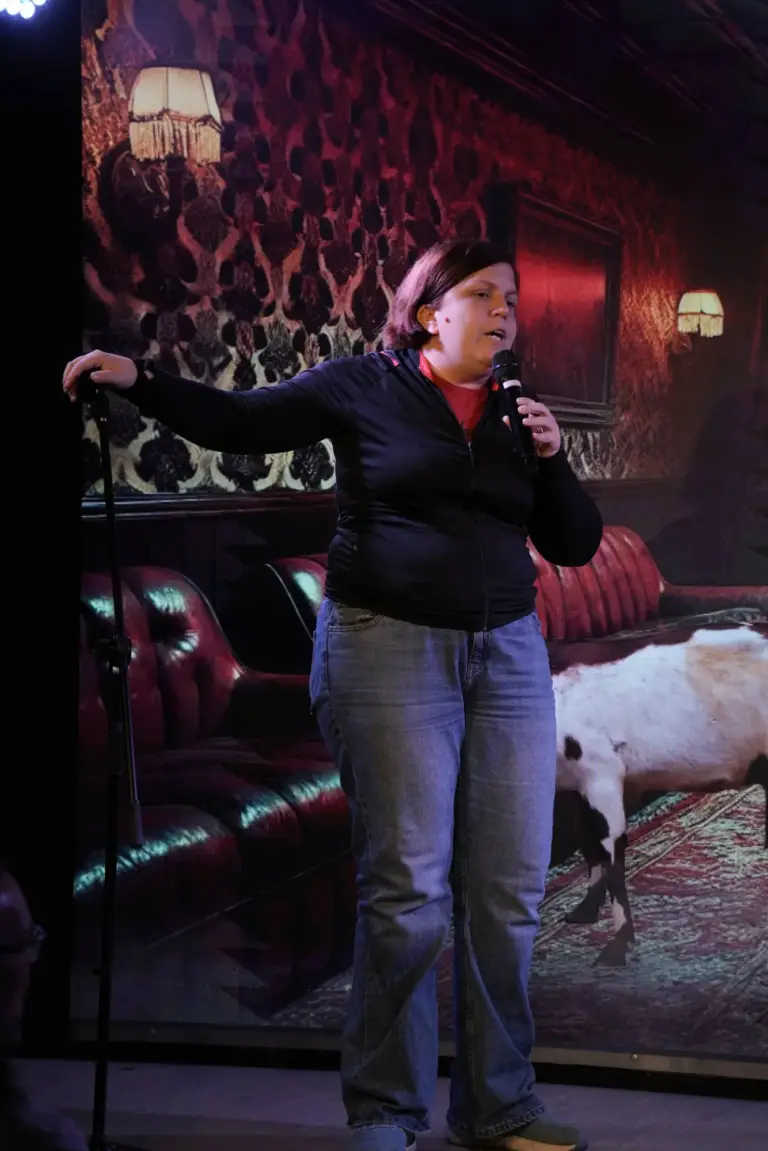 Artista con chaqueta oscura gesticulando en el escenario delante de unos sofás de terciopelo rojo