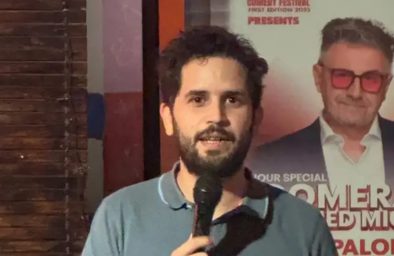 Cómico con barba sujetando el micrófono junto a un cartel de un espectáculo del festival