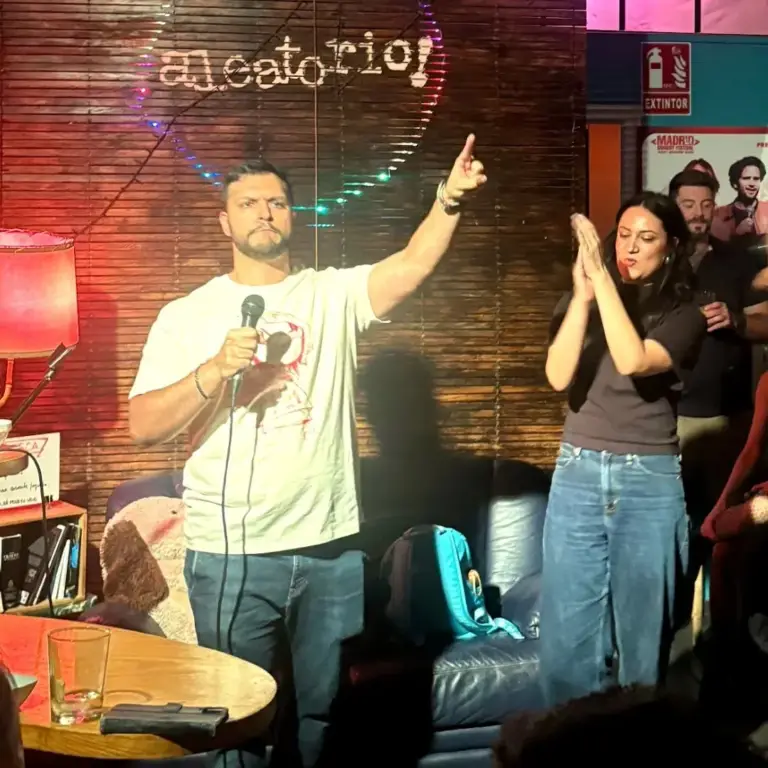 Cómico con camiseta blanca señalando hacia arriba mientras una compañera aplaude, en Aleatorio