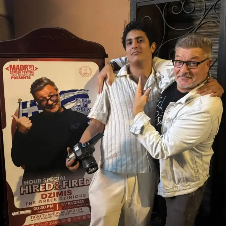Dos artistas posando junto a un cartel de un espectáculo del festival, uno con una cámara