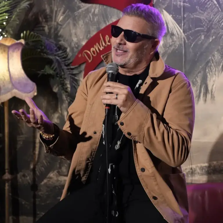 Cómico con gafas de sol y chaqueta camel actuando con una mano extendida