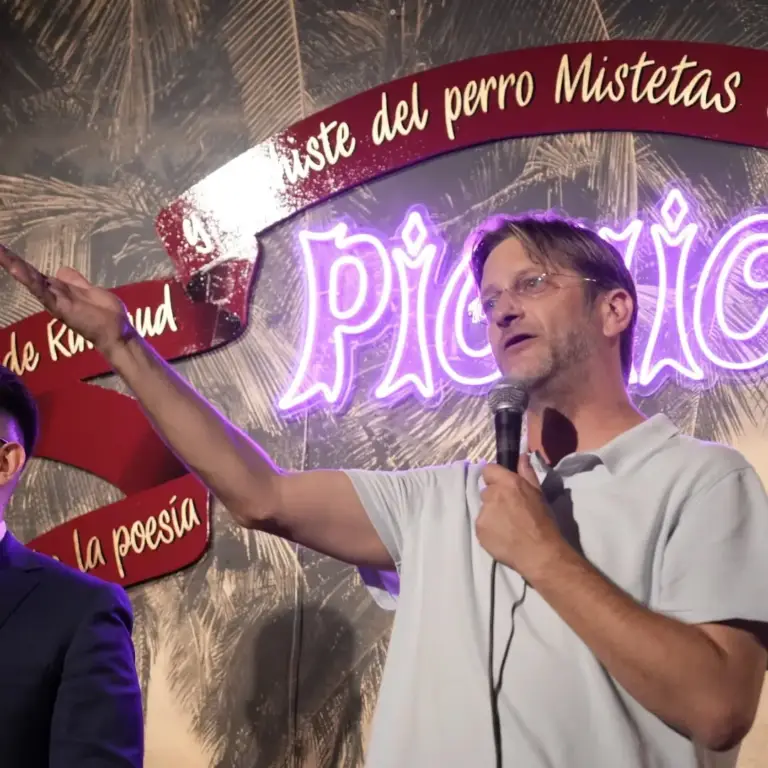 Cómico con polo gris señalando el rótulo de neón de Picnic mientras actúa