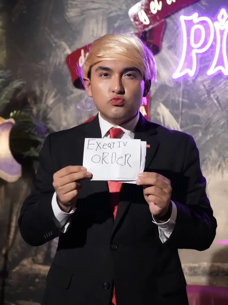 Artista con peluca rubia y traje enseñando un cartel escrito a mano que pone «Executive Order»
