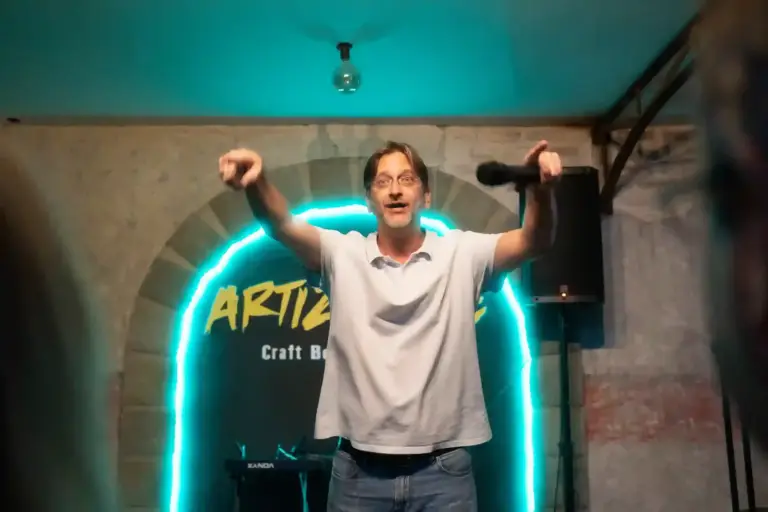 Cómico con los dos brazos en alto delante del arco de neón de Artizanale