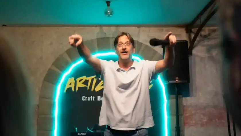 Cómico con los dos brazos en alto delante del arco de neón de Artizanale
