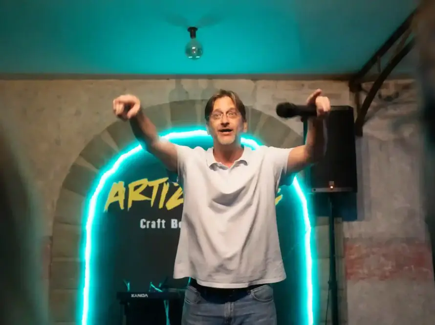 Cómico con los dos brazos en alto delante del arco de neón de Artizanale