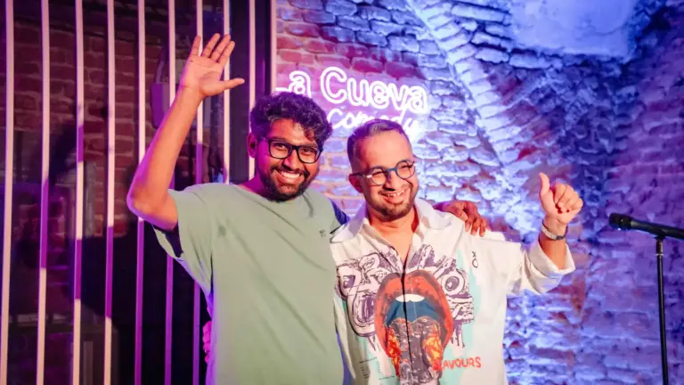 Dos cómicos saludando en el escenario bajo el rótulo de neón de La Cueva Comedy