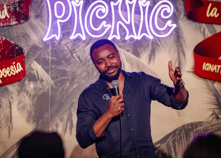 Cómico con camisa oscura gesticulando en mitad de un chiste bajo el rótulo de neón de Picnic