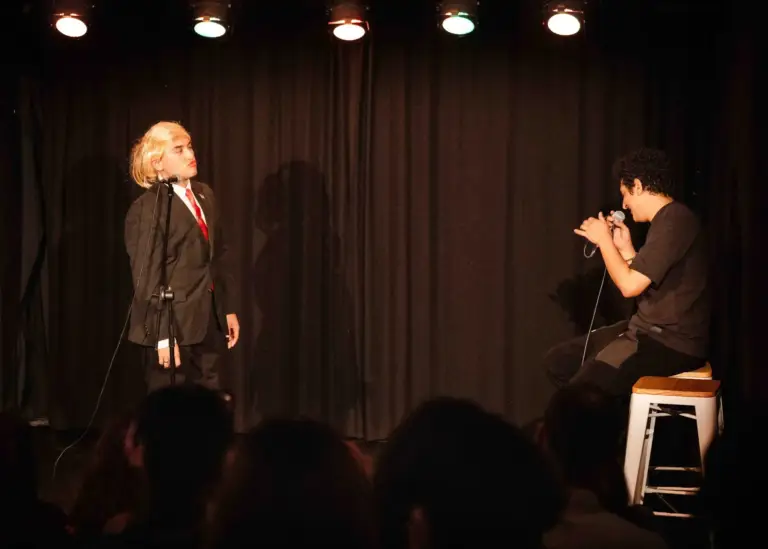 Artista con traje y peluca rubia en el escenario, con otro cómico sentado en un taburete