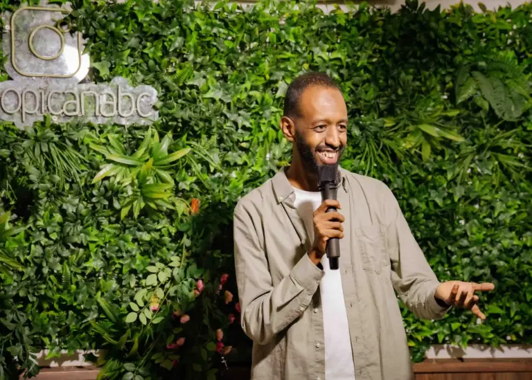 Cómico con sobrecamisa beis actuando delante de la pared de plantas de Tropicana